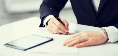 Homem branco com uma caneta na mão, tomando notas enquanto está com um vídeo aberto no ipad sobre a mesa